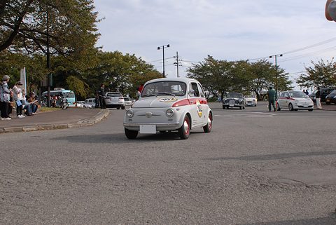 A small white two-door Fiat with a red stripe and rally plates crossing a car park, spectators at the edge. Opens a larger view