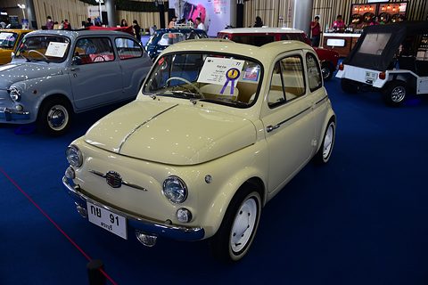 Cream Fiat 500 with a prize rosette in its windscreen, on blue carpet in a hall of classic cars. Opens a larger view