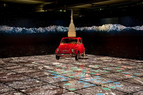 Small red Fiat 500 on a floor printed with a city map, a tall pale tower model rising behind it. Opens a larger view