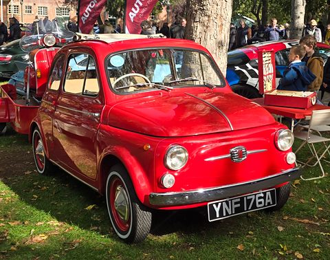 Bright red Fiat 500 with whitewall tyres and a chrome moustache badge, parked on grass under a tree among visitors. Opens a larger view
