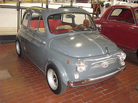 Grey Fiat 500 with red seats, on a brick floor beside a maroon car, a wire fence behind.