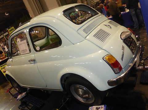 Cream Fiat 500 seen from high at the rear right, on a show stand with traders and visitors behind. Opens a larger view