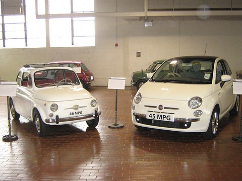 Small white classic Fiat 500 next to a larger white modern Fiat 500, both on a brick floor.