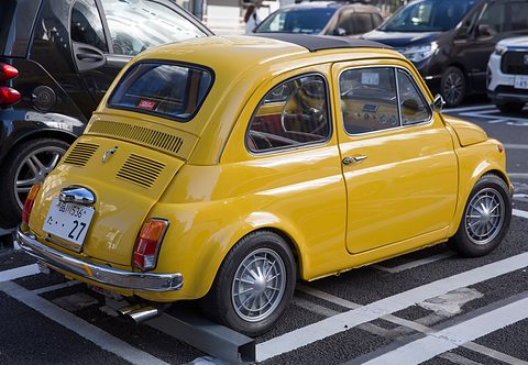 Yellow Fiat 500 from the rear left with alloy wheels and a white Japanese plate, between modern cars. Opens a larger view