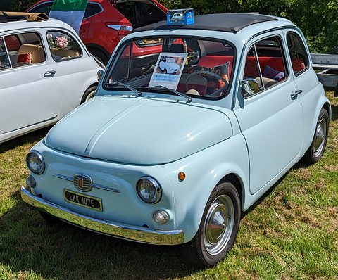 Pale blue Fiat 500 with a brochure on its dashboard and red seats, parked on grass among other cars. Opens a larger view