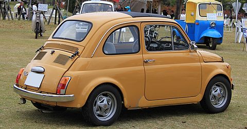 Ochre Fiat 500 from the rear left with alloy wheels and a louvred engine lid, a blue three-wheeled van behind. Opens a larger view