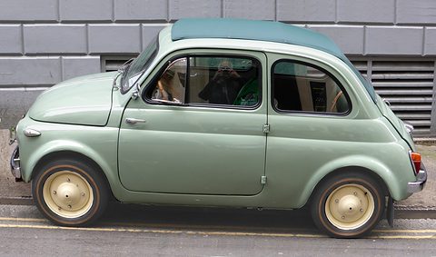 Side view of a pale green Fiat 500 with cream wheels and a teal roof, parked by a grey stone wall.