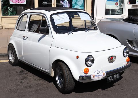 White Fiat 500 with a rally plate on its bumper and notes on the windscreen, parked before a shop window. Opens a larger view