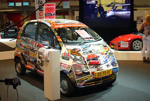 Small hatchback wrapped in a collage of car-magazine covers on a show stand, a red sports car behind. Opens a larger view