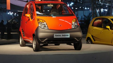 Front of an orange small hatchback with a black bumper and a Tata Nano plate, a yellow one beside it on a dark stand. Opens a larger view