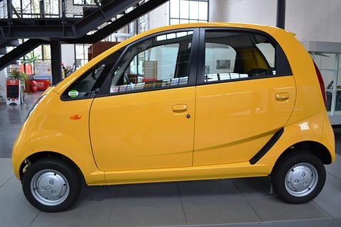 Side view of a bright yellow small hatchback in a hall with tall windows and steel stairs. Opens a larger view