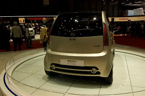 Rear of a champagne-coloured small hatchback with a Nano Europa plate on a white turntable, visitors behind. Opens a larger view