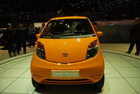 Front of an orange small hatchback with a Tata Nano plate on a white turntable, visitors around it. Opens a larger view