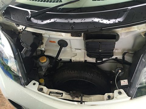 Open front of a white small car showing a spare wheel, fluid reservoirs and a battery, with no engine. Opens a larger view