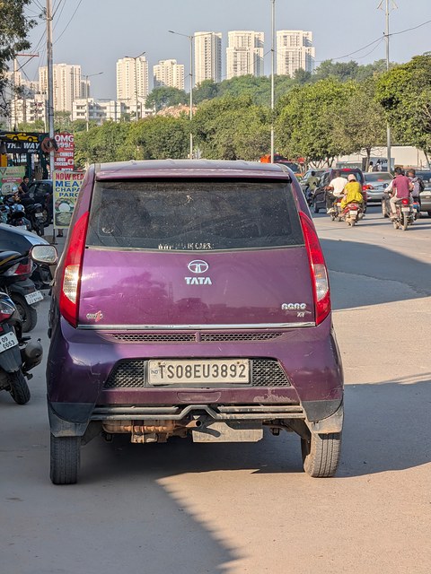 Rear of a purple small hatchback on a busy road with motorcycles, tower blocks on the skyline. Opens a larger view