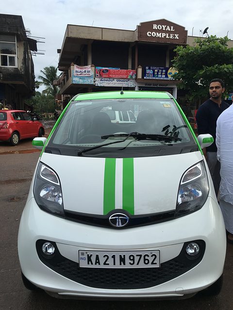 Front of a white small hatchback with twin green bonnet stripes and a Karnataka plate, people and shops behind. Opens a larger view