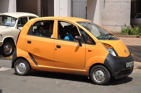 Side view of an orange small hatchback with passengers inside, a white Ambassador car behind. Opens a larger view