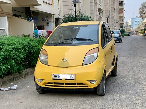 Front of a yellow small hatchback with a partly blanked plate, parked on a housing-estate road by a hedge. Opens a larger view