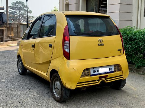 Rear of a dented yellow small hatchback with a blanked plate, parked on a housing-estate road. Opens a larger view