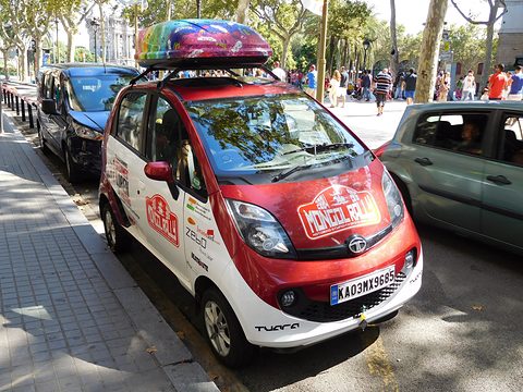 Red and white small hatchback with rally stickers and a rainbow roof box, parked on a tree-lined street. Opens a larger view