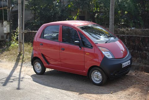 Red small hatchback with a black lower front parked on dusty ground beside a low wall and trees. Opens a larger view