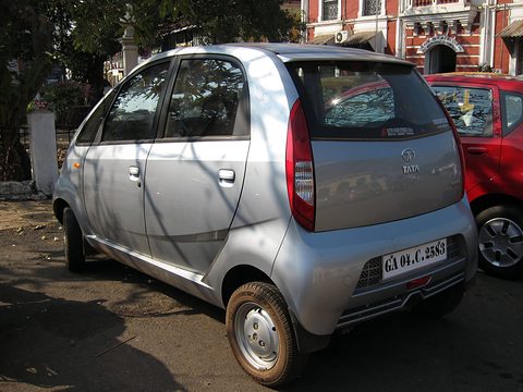 Rear of a silver small hatchback with a fixed rear panel and tall tail lamps, parked under trees by a red car. Opens a larger view