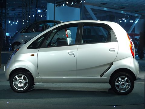 Side view of a silver tall-roofed small hatchback with alloy wheels on a dark show floor, another Nano behind. Opens a larger view