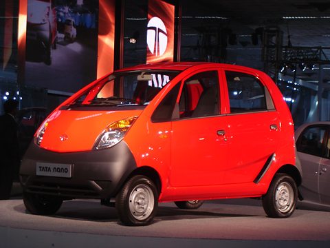 Red small hatchback with plain steel wheels and a black lower front on a show stand with a large Tata logo. Opens a larger view