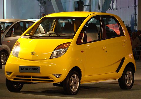Bright yellow small hatchback with a black Tata Nano plate on a show floor, a silver Nano behind. Opens a larger view