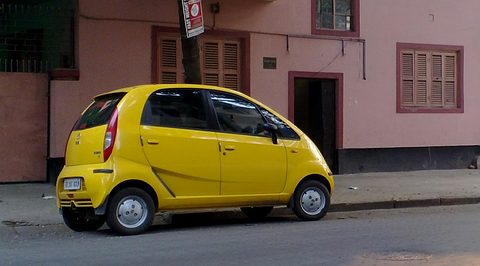 Side view of a bright yellow small hatchback on a street in front of a pink house with shuttered windows. Opens a larger view