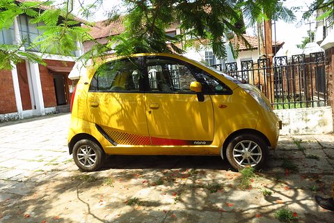 Side view of a bright yellow small hatchback with a red side stripe, in dappled shade by a house. Opens a larger view
