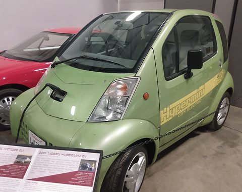 Pale green tall two-seat electric car with Hypermini script on its side and a charging lead, beside a sign board. Opens a larger view