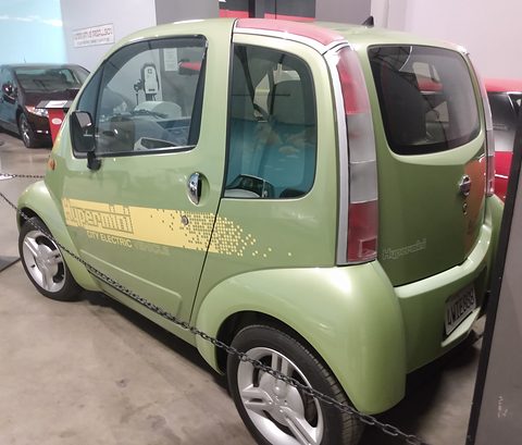 Rear of a pale green tall two-seat car with cream pixel graphics and a tall tail lamp, behind a chain. Opens a larger view