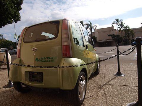 Rear of a pale green tall, rounded two-seat electric car with EV lettering behind chain barriers under palms. Opens a larger view