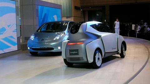 Narrow white tandem concept seen from behind next to a pale blue hatchback on a curved stand. Opens a larger view