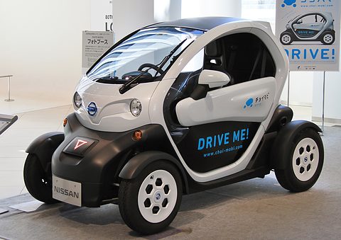 White and black tandem two-seater with Nissan badges and DRIVE ME! lettering on a white gallery floor. Opens a larger view
