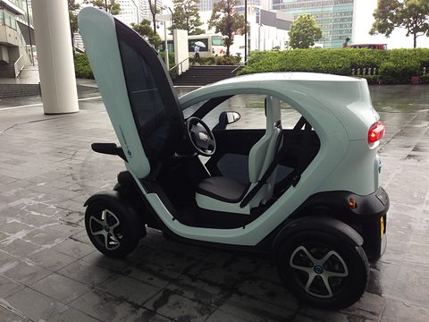Pale green tandem two-seat electric quadricycle with its scissor door up on a wet paved forecourt. Opens a larger view