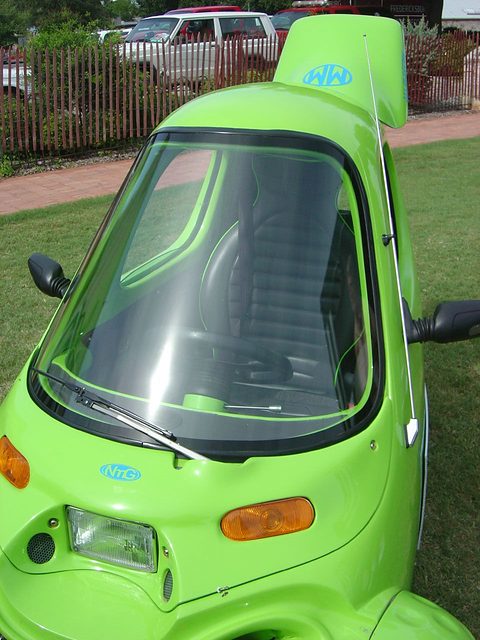 Bright green single-seater seen from the front with its door open, headlamps and amber indicators, on grass. Opens a larger view