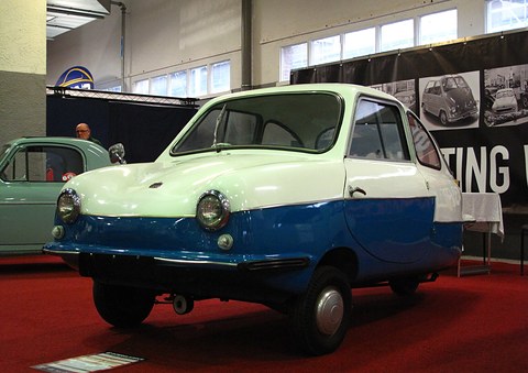 Two-tone pale green and blue three-wheeled microcar on red carpet at an indoor show. Opens a larger view