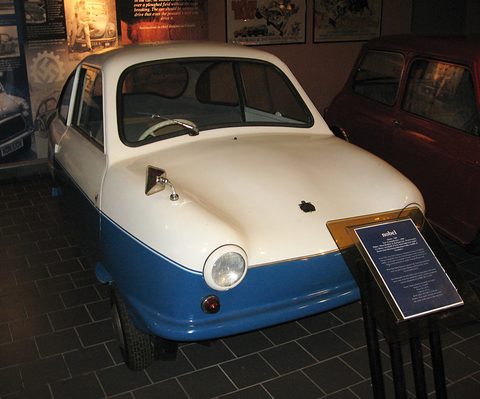 White and blue microcar facing the camera next to a museum information stand on a tiled floor. Opens a larger view