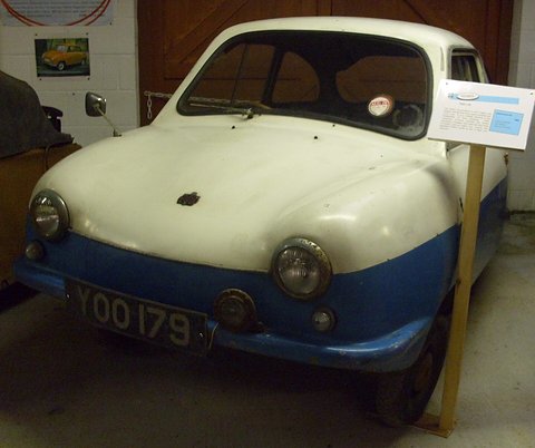 White and blue microcar beside an orange car in a museum, an information card at right. Opens a larger view