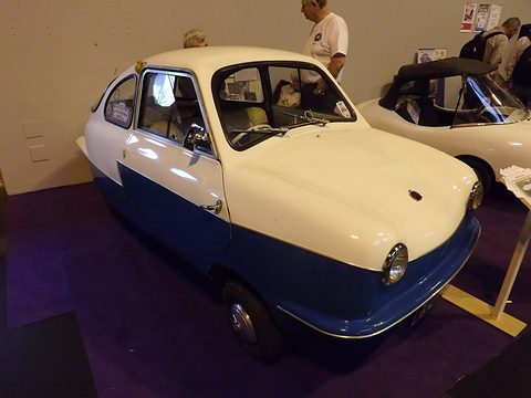 White and blue microcar with open-look windscreen on purple carpet beside a white sports car. Opens a larger view