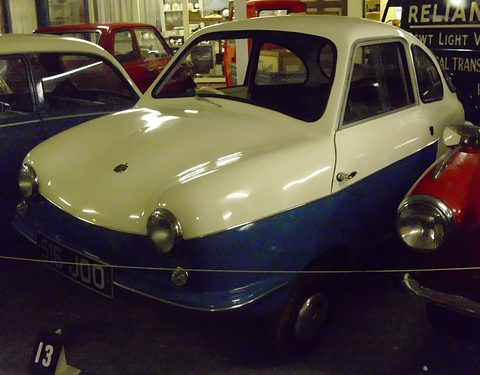 Cream and blue microcar with sloping bonnet behind a rope barrier, stand number 13 below. Opens a larger view