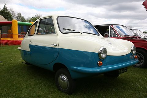White over blue microcar on a lawn, a bouncy castle and red car behind under a cloudy sky. Opens a larger view