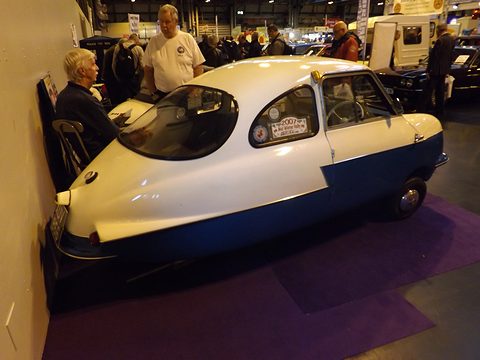 White over blue microcar with bubble rear window, a seated man at a table behind it. Opens a larger view
