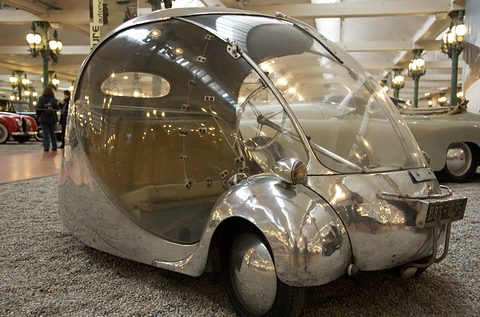 Low view of a gleaming egg-shaped car with a clear bubble roof under museum lamps.