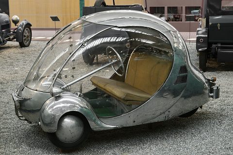 Polished aluminium egg-shaped car with a clear bubble, tan bench and spoked steering wheel. Opens a larger view