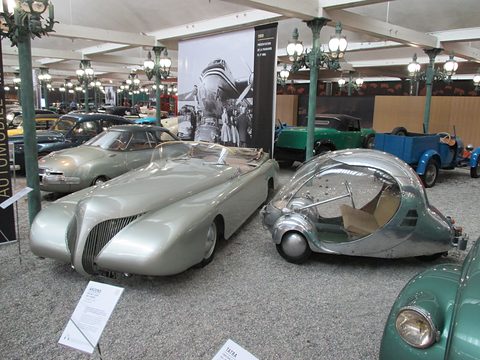 A long pale streamlined roadster beside a small aluminium egg car on gravel, a photo panel behind.
