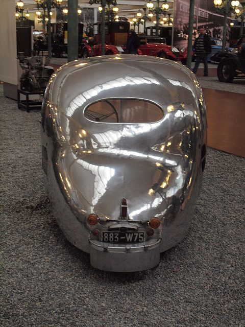 Rear of a mirror-polished aluminium egg-shaped car with a small oval window and a number plate. Opens a larger view