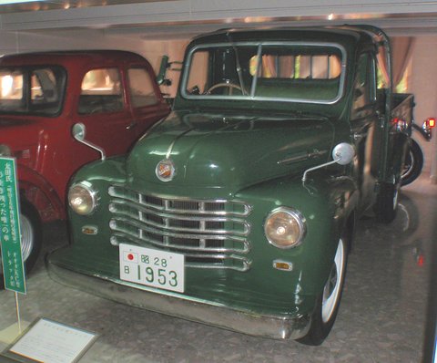 Dark green small pick-up with a chrome grille and plate reading 1953, behind glass, a red saloon beside it. Opens a larger view